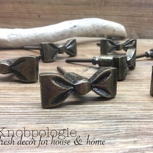 May include: A collection of antique bronze bow-shaped cabinet knobs. The knobs are arranged on a wooden surface, with a driftwood piece in the background. The text "Knobpologie fresh decor for house & home" is visible.