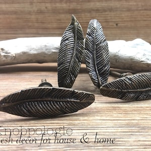Peut inclure: Quatre boutons de meuble en forme de plume de couleur bronze, disposés sur une surface en bois. Les boutons présentent des veines détaillées et sont destinés à la décoration intérieure. Le texte "Knobpologie fresh decor for house & home" est également visible.