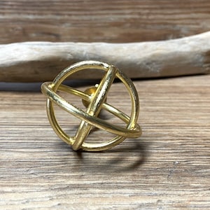 May include: A gold-colored, spherical decorative object with intersecting circular bands. The orb-like design is set against a wooden background, creating a rustic aesthetic. The object's metallic finish adds a touch of elegance to the piece.