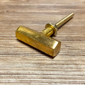 Op de afbeelding: Een goudkleurige kastknop met een zeshoekige handgreep en een cilindrische steel. De knop is gemaakt van metaal en heeft een glanzende afwerking. De knop is ontworpen om aan een kastdeur of lade te worden bevestigd.
