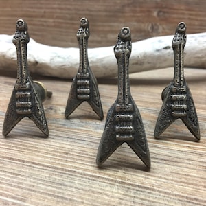 May include: Four bronze-colored guitar-shaped cabinet knobs arranged on a wooden surface. The knobs are detailed with strings and tuning pegs, and are designed in a V-shape. The knobs are likely for a cabinet or drawer.