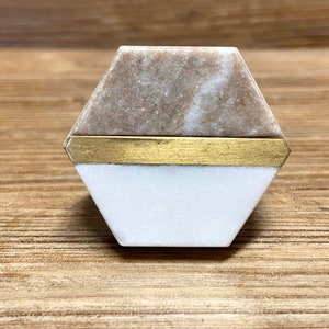 May include: A hexagonal cabinet knob with a two-tone design. The top section is a light brown marble, the bottom is white marble, and a gold-colored metal band separates the two. The knob is set against a wooden surface.