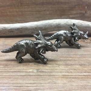 Op de afbeelding: Twee zilverkleurige dinosaurusbeeldjes, mogelijk triceratops, met een gestructureerd oppervlak. De dinosaurussen zijn naar rechts in de afbeelding gericht en rennen.
