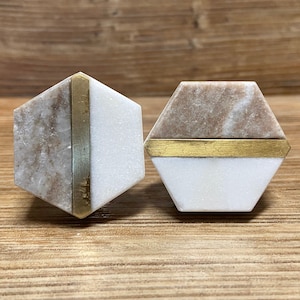 May include: Two hexagonal cabinet knobs. Each knob features a combination of white and brown marble with a brass accent. The knobs are designed for furniture and home decor projects.