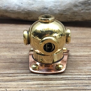 May include: A miniature brass diving helmet with a copper base, displayed on a wooden surface. The helmet features a round viewing port and is a decorative item. The logo "Knobpologie fresh decor for house & home" is visible in the lower left corner.