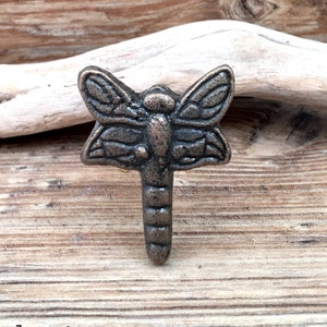 Könnte beinhalten: Ein Metall-Libellenhaken in antiker Bronze-Optik. Der Haken ist auf einem Stück Treibholz montiert.