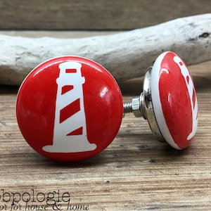 Puede incluir: Dos pomos de armario rojos y blancos con temática de faro. Los pomos tienen forma redonda con un diseño de faro blanco sobre fondo rojo. Los pomos están sujetos a una base de metal plateado. El texto "Knobpologie fresh decor for house & home" está en la parte inferior.