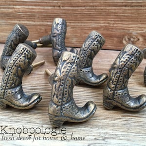 Peut inclure: Boutons de meubles en bronze en forme de bottes de cowboy. Les boutons présentent des détails complexes et sont disposés sur une surface en bois. Le texte "Knobpologie fresh decor for house & home" est visible.