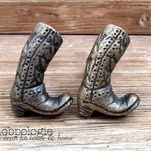Peut inclure: Deux boutons de meuble en forme de bottes de cowboy de couleur bronze. Les boutons présentent des coutures détaillées et une finition vieillie. Les bottes sont inclinées, suggérant un élément de design décoratif pour la décoration intérieure.