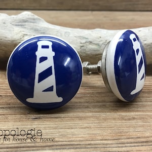 Puede incluir: Dos pomos de armario azules y blancos con temática de faro. Cada pomo presenta un diseño de faro blanco sobre un fondo azul. Los pomos son redondos con un borde blanco y un tornillo plateado.
