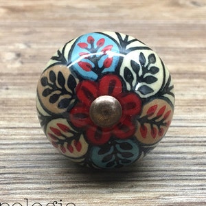 May include: A decorative ceramic cabinet knob with a floral design. The knob features a red flower at the center, surrounded by blue, beige, and black leaf patterns. The knob has a bronze-colored screw and the text "Knobpologie fresh decor for house & home".