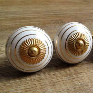 May include: Four white cabinet knobs with gold accents. Each knob features a gold sunburst design and gold stripes. The knobs are arranged in a row on a wooden surface. The text "knobpologie" is visible.