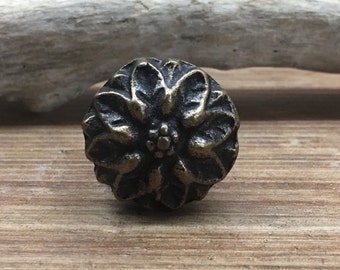 May include: A round, ornate, brass-colored cabinet knob with a floral design. The knob is on a wooden surface.