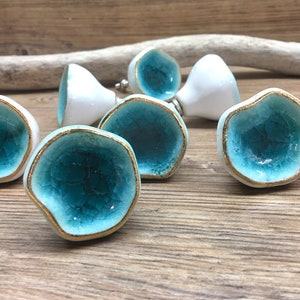 May include: A collection of ceramic cabinet knobs with a unique design. Each knob features a turquoise crackle glaze center, a gold-colored rim, and a white base. The knobs are irregularly shaped and add a decorative touch to furniture.
