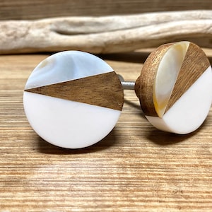 May include: Two decorative cabinet knobs. Each knob features a white, wood, and pearlescent design. The knobs are round with a metal screw for installation. The wood is a medium brown color. The knobs are on a wooden surface.