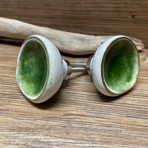 May include: Two white ceramic cabinet knobs with a green interior and gold trim. The knobs are mounted on silver hardware and are set against a wooden background. These decorative knobs are ideal for adding a touch of elegance to furniture.