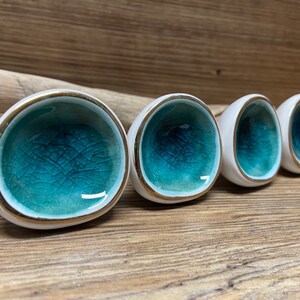 May include: Four white ceramic cabinet knobs with a teal crackle glaze interior and gold trim. The knobs are arranged in a row, showcasing their unique design and craftsmanship. These decorative knobs add a touch of elegance to any furniture piece.