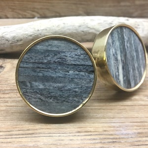 May include: Two round cabinet knobs with a brass finish and a gray and white stone center. The knobs are set against a wooden background. The knobs are a decorative hardware item.