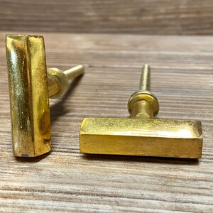 Op de afbeelding: Twee messingkleurige kastgrepen met een T-bar ontwerp. De grepen hebben een rechthoekige balk en een cilindrische steel. De hardware is geplaatst tegen een houten achtergrond. Deze grepen zijn geschikt voor laden, kasten en ander meubilair.