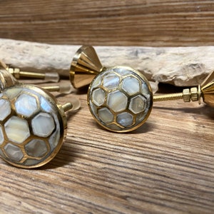 May include: Set of four decorative cabinet knobs. Each knob features a round, white and gold honeycomb design. The knobs have a brass finish and are mounted on threaded rods with nuts.