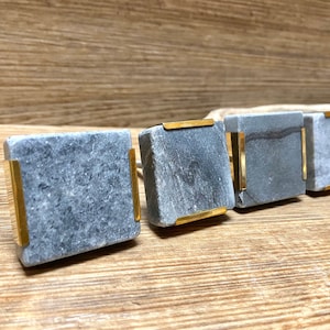 May include: Four square cabinet knobs made of gray stone with gold-colored metal accents. The knobs are arranged in a row on a wooden surface, showcasing their design and texture. Each knob is approximately 1.5 inches square.