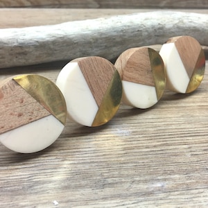 以下が含まれることがあります： モダンなデザインの丸いキャビネットノブ4つ。各ノブは、明るい木材、白、金色の金属の組み合わせが特徴です。ノブは木製の表面に一列に並んでおり、幾何学模様を示しています。