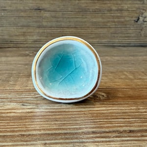 Puede incluir: Un pequeño plato redondo de cerámica con interior turquesa y borde dorado. El plato tiene un exterior blanco y está sobre una superficie de madera. El interior turquesa tiene un efecto craquelado.