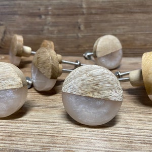Könnte beinhalten: Eine Sammlung runder Schrankknöpfe in zweifarbigem Design. Jeder Knopf besteht aus einer oberen Hälfte aus hellem Naturholz und einer unteren Hälfte aus glattem, halbtransparentem, perlmuttfarbenem Harz, befestigt an einem Metallgewindebolzen.