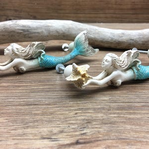 May include: Two mermaid-shaped cabinet knobs with turquoise tails and seashell accents. The mermaids are holding seashells in their hands.