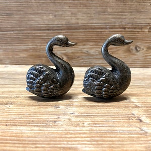 May include: Two small, silver-colored swan figurines. The swans are facing the right side of the image and are positioned on a wooden surface.