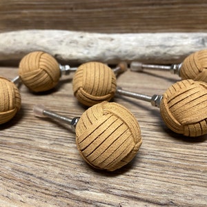 Pode incluir: Um conjunto de sete puxadores de armário em couro castanho. Cada puxador, de forma esférica e com um design entrançado, está fixado a um parafuso metálico. Dispostos sobre uma superfície de madeira, para decoração.