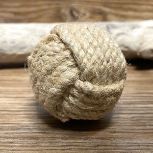 May include: A decorative ball made of tightly woven, natural-colored rope. The ball is round and knotted, with visible texture and detail. The background is a wooden surface.