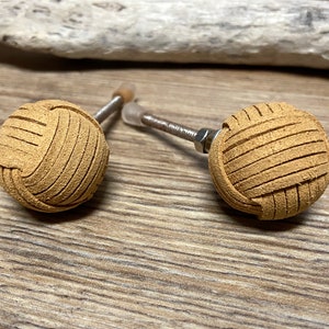 May include: Two tan leather cabinet knobs with a woven design. Each knob is spherical and attached to a metal rod with a screw. The knobs are on a wooden surface.