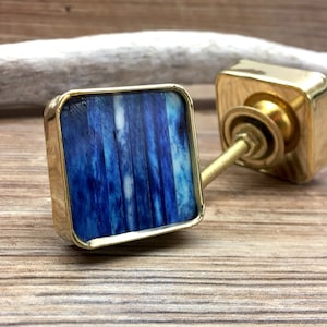 Puede incluir: Dos tiradores cuadrados para armario con acabado dorado e incrustaciones azules y blancas. Los tiradores tienen un diseño rectangular con borde dorado. La ferretería está sobre un fondo de madera.