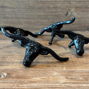 May include: Three black metal bull head cabinet pulls. The pulls are in a rustic style, with a glossy finish. The bull heads are detailed with horns and facial features. They are mounted on a wooden surface.