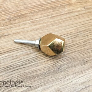 May include: A gold-colored, faceted cabinet knob with a silver screw. The knob has a geometric design and is set against a light brown wood grain surface. The text "knobpologie fresh decor for house & home" is visible.