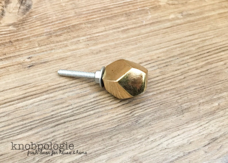 May include: A gold-colored, faceted cabinet knob with a silver screw. The knob has a geometric design and is set against a light brown wood grain surface. The text "knobpologie fresh decor for house & home" is visible.
