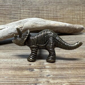 Op de afbeelding: Een bronzen metalen triceratops dinosaurus beeldje. Het beeldje is gedetailleerd met getextureerde schubben en heeft een lange staart.