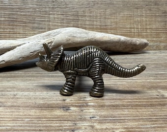 Puede incluir: Una figurita de dinosaurio triceratops de metal de color bronce. La figurita está detallada con escamas texturizadas y tiene una cola larga.