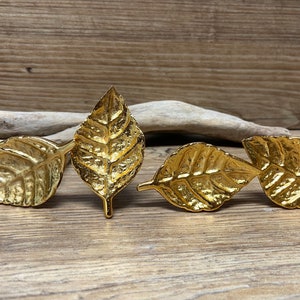 May include: Four gold-colored leaf-shaped decorative objects. The leaves have detailed veining and a metallic finish. They are arranged on a wooden surface, suggesting a nature-inspired theme.