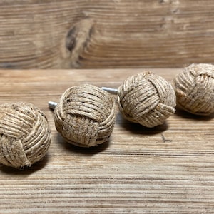 Op de afbeelding: Vier decoratieve kastknoppen. Elke knop is een bol van strak gewikkeld natuurlijk gekleurd touw. De knoppen hebben een zilverkleurige metalen steel voor montage. De knoppen zijn gerangschikt op een houten oppervlak.
