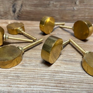 May include: A collection of seven brass cabinet knobs with a flat, rounded design. Each knob has a threaded post for easy installation. The knobs are arranged on a light brown wooden surface.