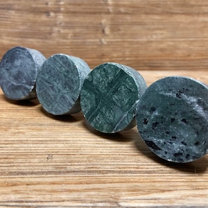 Può includere: Quattro pomelli circolari in pietra disposti in linea diagonale. Ogni pomello presenta un motivo naturale unico nei toni del grigio scuro, antracite e verde bosco profondo con venature minerali visibili e una finitura liscia e lucida.