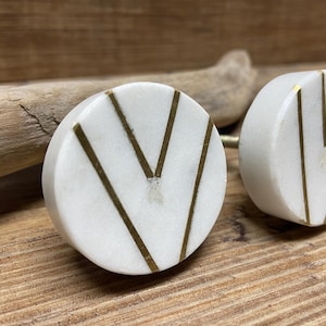 May include: Two white marble cabinet knobs with gold-colored chevron designs. The knobs are round and have a brass stem. The background is a wooden surface with a piece of driftwood.