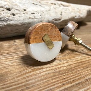 May include: Two decorative cabinet knobs. Each knob features a half-circle of light-colored wood and a half-circle of white stone, joined by a brass accent. The knobs have a brass screw and are set against a wooden surface.