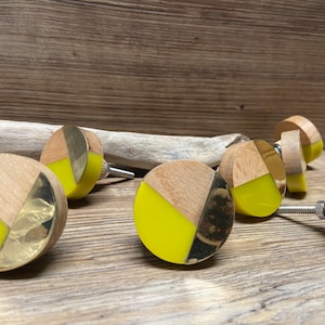 May include: A selection of cabinet knobs with a decorative design. The knobs combine light wood, brass, and bright yellow resin. Each round knob has a metal screw for fitting. The knobs are displayed on a wooden surface.