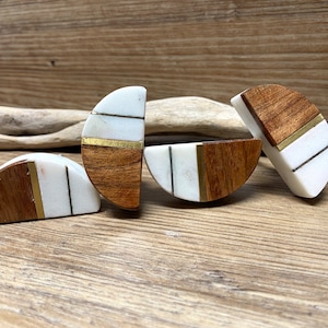 Może przedstawiać: Cztery uchwyty do szafek w kształcie półksiężyca. Każdy uchwyt łączy biały marmur, ciepłe drewno i mosiężne akcenty. Uchwyty są ułożone na drewnianej powierzchni, podkreślając ich design i fakturę. Mają około 5 cm długości.