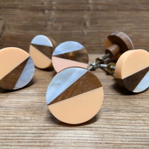 Pode incluir: Um conjunto de seis puxadores decorativos para armários. Cada puxador apresenta um design circular com uma secção pêssego, uma secção de madeira escura e uma secção perolada. Os puxadores estão sobre um fundo de madeira.