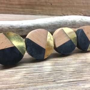 May include: Four round wooden cabinet knobs with a geometric design. Each knob features a natural wood section, a black section, and a gold-colored metal section. The knobs are arranged in a row, showcasing their decorative appeal.