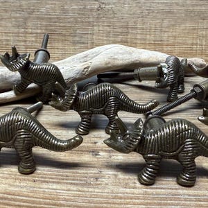 Op de afbeelding: Vijf antieke bronzen knoppen in de vorm van een dinosaurus met een gestructureerd oppervlak. De knoppen bevinden zich in verschillende poses, waaronder staand, lopend en rennend.